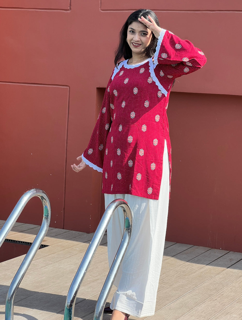 Woman wearing a red and white patterned top with white pants standing against a brown wall.